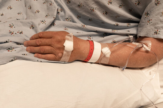 Arm and hand of a man in a hospital gown and bed, IV lines, wristbands, bandages, healthcare creative copy space, horizontal aspect