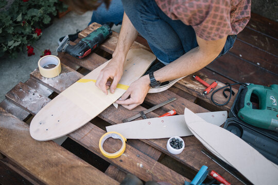 Cropped man sticking paper tape to skateboard in yard. Male hands sticking tape to wooden board for painting outdoor.