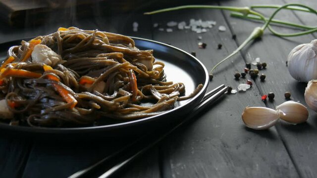 Freshly boiled steam asian buckwheat noodles soba with vegetables onion pods, carrots, paprika and pieces of chicken, on a stylish black plate and Chinese chopsticks.