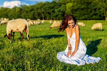 Beautiful woman wearing white dress enjoys on meadows with sheep in the background.