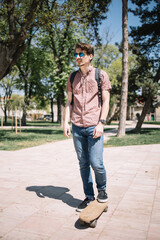 Cool guy with sunglasses posing with skateboard. Portrait of man standing in park with skateboard during sunny day.