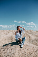 Fototapeta na wymiar Woman in blue jeans, white shirt and a hat seatting on rocks. White hills of refuse pile and blue sky. Woman in a desert on spoil heap.