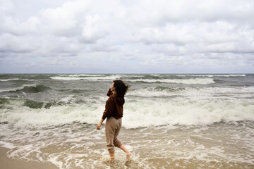 young woman walking on the beach with her feet in the water, watching tide on a Baltic Sea, windy day with waves, day on a shore, enjoying nature, relaxing outside by seaside 