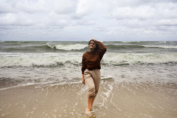 young woman walking on the beach, young woman standing with her feet in the water, watching tide, windy day, outdoor activities