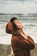 young woman on beach, tide at a Baltic Sea, young woman in hoodie wearing hood, enjoying the sound of the sea, listening to the waves, windy day, relaxing