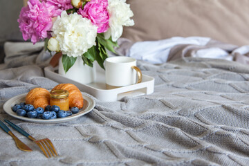 Cozy breakfast in bed, cup of coffee, blueberries, jam and croissants on grey bed.  Pink and white pions in the vase.
