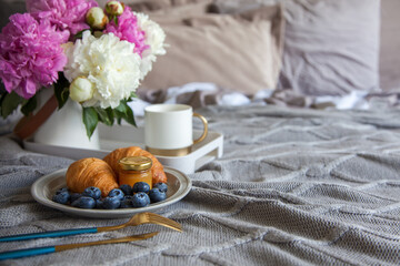 Cozy breakfast in bed, cup of coffee, blueberries, jam and croissants on grey bed.  Pink and white pions in the vase.