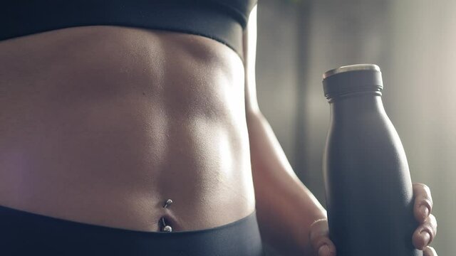 Backlit close up of fitness woman abs with hand keeping sport bottle. Athlete woman abs muscles detail. Abs muscular build achievement.