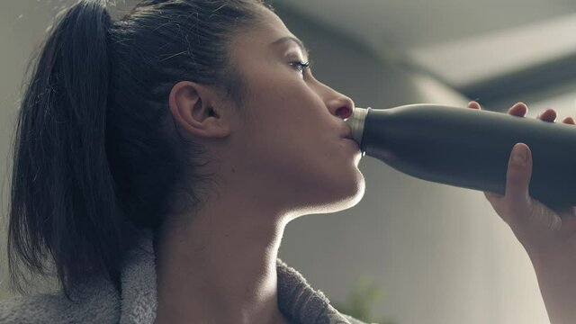 fitness woman drinking from sport bottle during a workout pause at home or gym. Close backlit portrait of active woman drinking from water bottle while resting from training