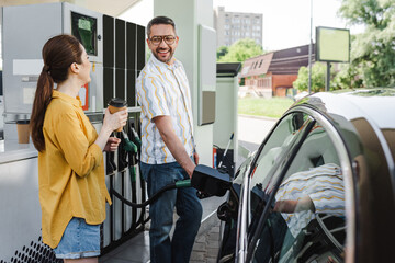 Selective focus of smiling man looking at wife with coffee to go while refueling car on gas station