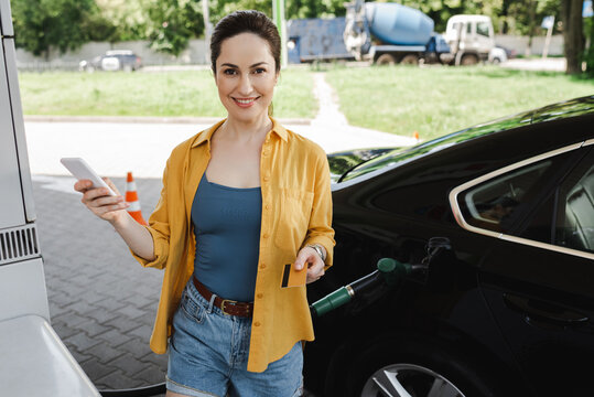 Smiling Woman Holding Credit Card And Smartphone Near Car On Gas Station