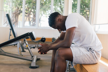 Desperate African American male athlete sitting on bench during break in fitness training in modern...