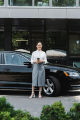 Selective focus of smiling businesswoman holding coffee to go near auto on urban street