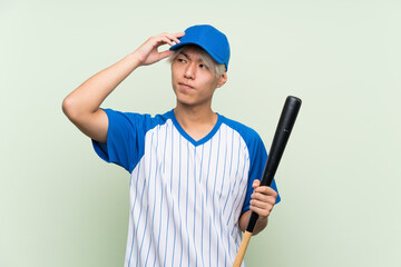 Young asian man playing baseball over isolated green background having doubts and with confuse face expression