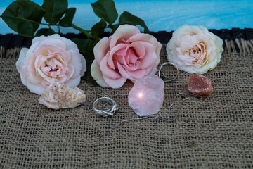 On jute fabric are semiprecious stones rose quartz, dolomite, halite. Nearby are silver rings and a chain. In the background there are three pink roses..
