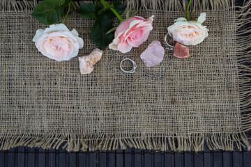 Top view. On the jute fabric are semi-precious stones pink quartz, dolomite, halite and three pink roses. Nearby are silver rings and a chain.