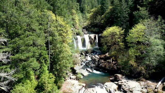 Drone footage flying straight up showing the water fall at Coquille falls and the pristine clean water flowing between rocks and boulders surrounded by a valley of trees.
