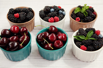 Fresh berries of cherries, raspberries, blackberries, blueberries, mint leaves, in bowls on a gray wooden background.