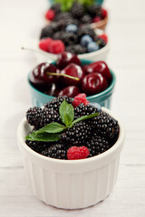 Fresh berries of cherries, raspberries, blackberries, blueberries, mint leaves, in bowls on a gray wooden background.