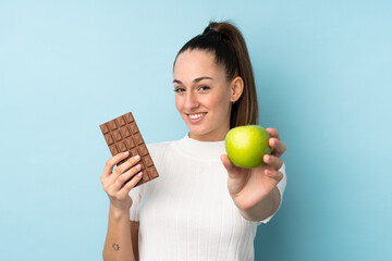 Young brunette woman over isolated blue background taking a chocolate tablet in one hand and an...
