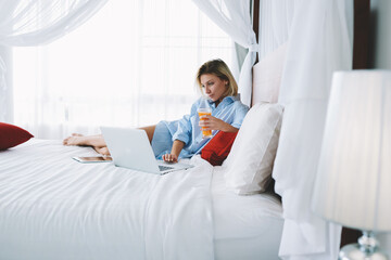 Young woman watching movie on laptop computer relaxing in bed with fresh juice in morning, female freelancer enjoying remote work using netbook connected to wifi for read news recreating in hotel room