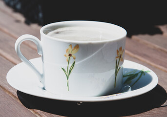 Biała filiżanka w kwiaty z kawą, lekka pianka, bez mleka. Naturalny, drewniany stół.