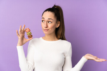 Young brunette woman over isolated purple background holding colorful French macarons and making doubts gesture