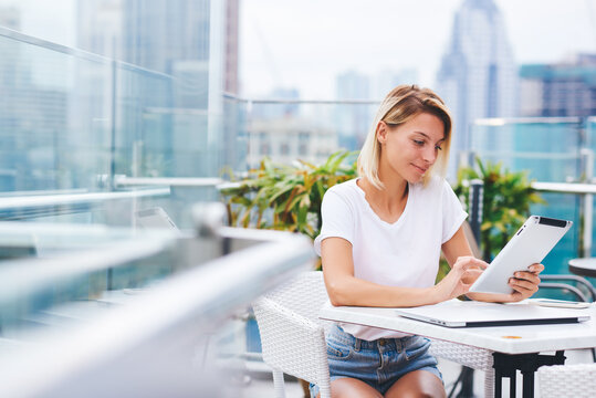 Concentrated female hipster girl spending time on hobby reading electronic book via modern tablet enjoying weekends sitting in skyscraper rooftop cafe with gorgeous panoramic and wifi connection