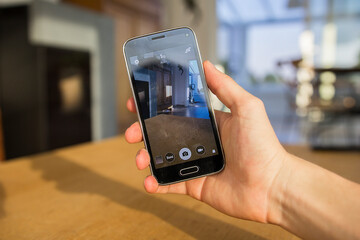 A hand holding a mobile phone which displays the camera app on the touch screen. The image is taken in the living room of a home.