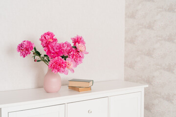 big bouquet of fuchsia peonies in a pink vase in a bright room are on a white chest of drawers, books are nearby. cozy house, fresh flowers at home, home greenhouse, minimal design, loft house