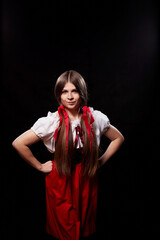 A girl in national ethnic clothing poses in a Studio on a black background. A young woman in a red dress and white shirt with long hair and ponytails in a dark room.
