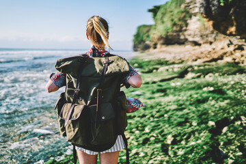 Fototapeta na wymiar Rear view of blonde young tourist with backpack strolling outdoors on coastline enjoying green hills and environment.Back view of hipster traveller with rucksackstanding on green shore washed by ocean