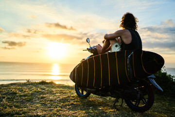 Back view of young sportive male tourist recreating outdoors watching at sunset near to ocean with ray reflection.Man surfer traveler relaxing outside sitting on cool motorcycle at evening time