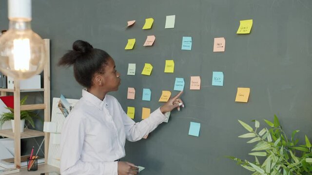 Female office worker is sticking bright colorful sticky notes on wall in workplace busy with project. Businesspeople, creative work and workspace concept.