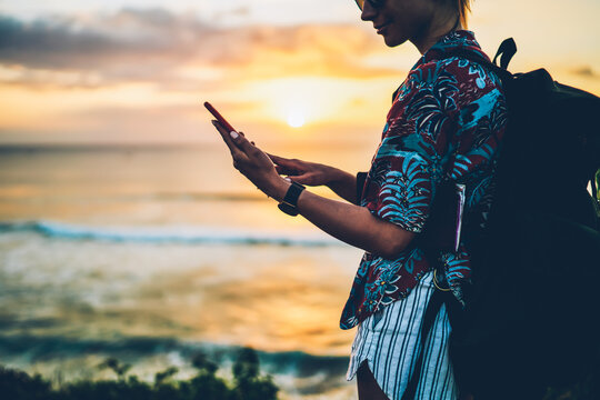 Cropped image of positive hipster girl dressed in casual clothing enjoying spending summer vacation against ocean landscapes.Young woman tourist with rucksack sending messages to friends via phone