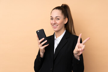 Fototapeta premium Young business woman using mobile phone over isolated background smiling and showing victory sign