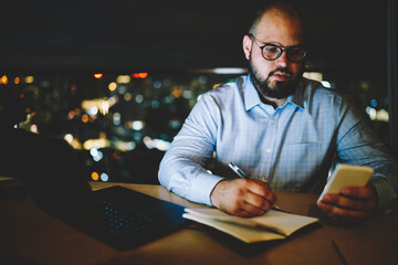 Concentrated businessman noting contact information from cellphone to notepad working...