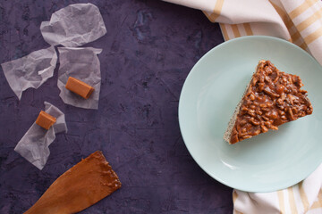 Piece of pie covered with caramel and suflower seeds in light blue plate with  wooden spatula and candies on a violet grunge background
