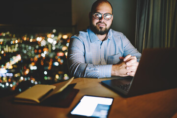 Portrait of prosperous businessman sitting at desktop in apartments with night city window view and modern technology, male freelancer looking at camera staying late for distance job online