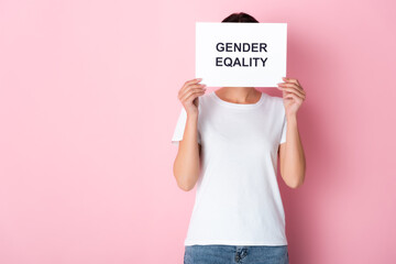 woman in white t-shirt covering face with gender equality lettering on placard and standing on pink