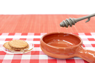 Cuchara de miel goteando en un tazón arcilla, sobre la mesa un mantel rojo y blanco y galletas