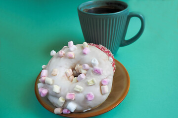 two doughnuts with pink icing and a white one with marshmallows on a brown round plate with coffee in a turquoise mug on a turquoise background side view