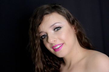 Naklejka premium close up portrait of beautiful brunette young girl happily smiling, looking at camera with fresh make up on black background, pink lipstick curly hair over bare, naked shoulders, studio shot,