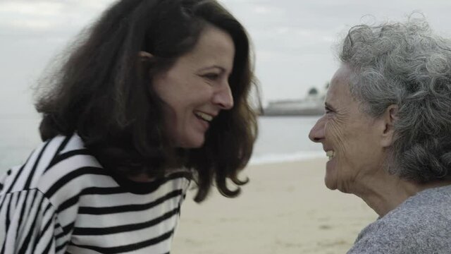 Cheerful adult daughter making silly faces and senior mother smiling, and laughing. Happy women leaning foreheads. Sea waves flowing on background. Cropped view. Family, seashore and weekend concept