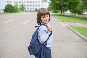 Fototapeta premium happy girl schoolgirl 8 years old European appearance with a backpack walks on the school yard in the afternoon on the street looking at the camera close-up
