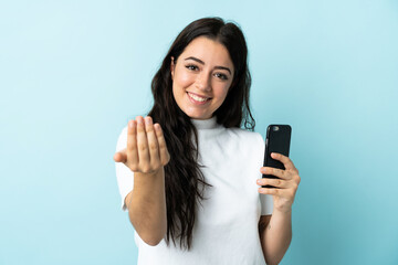 Young woman using mobile phone isolated on blue background inviting to come with hand. Happy that...