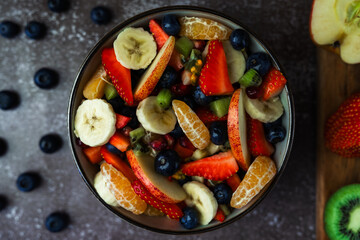 Overhead view of fruit salad in bowl with scattered blueberries and wooden chopping board with prepared fruits