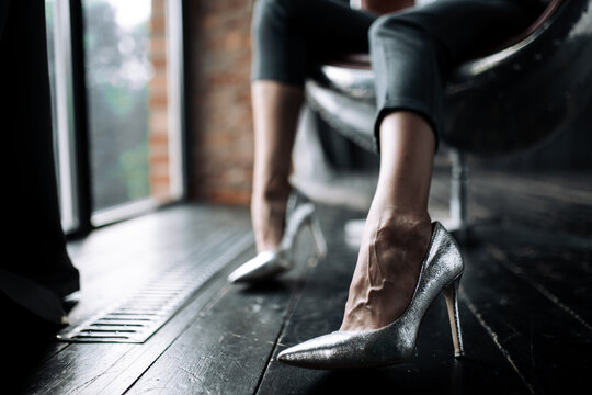 A close-up photograph of the legs of a girl in silver high heel shoes sitting in an aviator chair in a loft room.