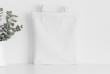 White tote bag mockup with a eucalyptus in a vase on a white table.