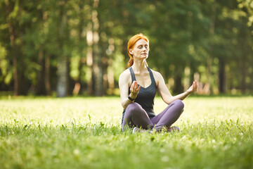 Serene relaxed mature redhead woman sitting with crossed legs on grass and meditating with closed...